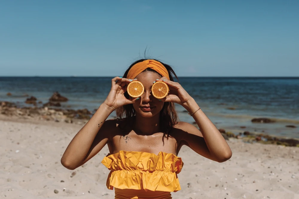 Ženska na plaži z rezinama pomaranče pred očmi simbolizira svežino in naravno moč aljaškega ribjega olja z omega-3.